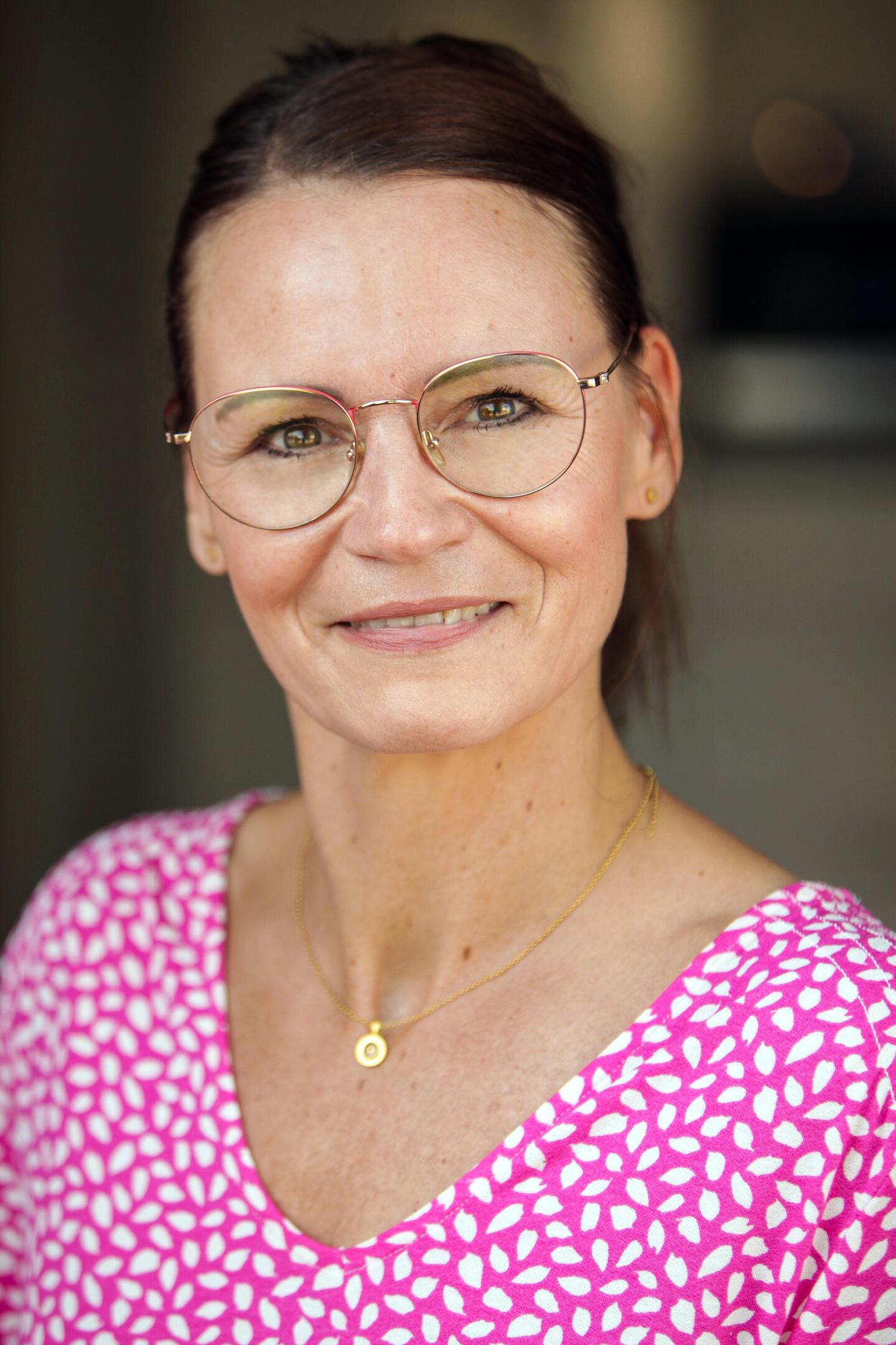 Eine Frau mit runden Brillengläsern und einem pinken Oberteil mit weißem Muster lächelt in die Kamera.